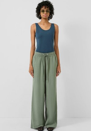 Vrouw met krullend haar die een zonnebril draagt, een blauw mouwloos topje, losse olijfgroene broek en zwarte schoenen, staand tegen een effen achtergrond.