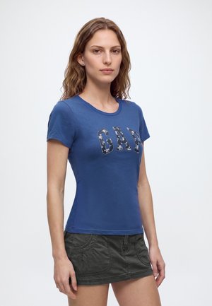 Jeune femme aux cheveux bruns ondulés portant un t-shirt bleu GAP et une mini-jupe gris foncé, debout devant un fond blanc uni.