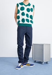 Maglione senza maniche verde e bianco con grandi pois, abbinato a pantaloni blu scuro e sneakers blu con strisce gialle, accanto a un carrello metallico.