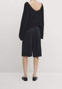 Maglione nero lavorato a maglia con schiena scoperta, abbinato a pantaloni corti scuri su misura e scarpe slip-on nere. Tessuto morbido, vestibilità comoda.
