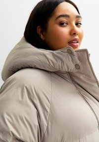 Mujer con cabello liso y oscuro vestida con una chaqueta acolchada beige de cuello alto, mirando por encima del hombro frente a un fondo claro y liso.