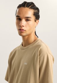 Béžové bavlněné tričko s kulatým výstřihem a krátkým rukávem, na hrudi s malým bílým logem "VANS". Měkká textura, ležérní design.