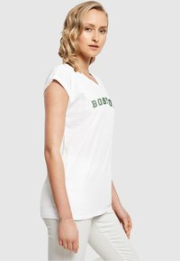 Weißes Baumwoll-T-Shirt mit grütem "BOSTON"-Text, kurzen aufgerollten Ärmeln, V-Ausschnitt und lockerem Schnitt. Getragen mit hellen Hosen.
