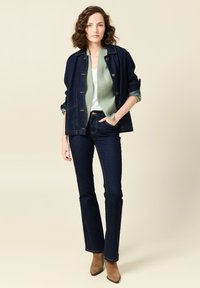 Giacca di jeans blu scuro, indossata sopra un maglione trapuntato verde chiaro, abbinata a jeans svasati scuri e stivaletti con tacco color nocciola.