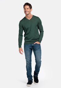 Groene V-hals trui van zachte stof, gecombineerd met slim-fit blauwe jeans en zwarte sneakers. Model staat in een ontspannen pose.