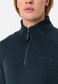 Pulover din fleece cu fermoar, de culoare albastru închis, cu un tricot texturat, având un guler înalt și un logo mic pe piept. Fermoar de culoare închisă, asortat.