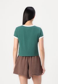 Camiseta corta verde de algodón, con un cuello redondo blanco y dobladillos en las mangas. Combinada con pantalones cortos marrones texturizados.