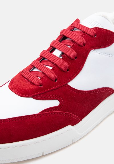 Λευκό αθλητικό παπούτσι με κόκκινα σουέτ πέτα και κορδόνια που ταιριάζουν σε λευκή λαστιχένια σόλα.