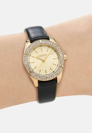 Gouden polshorloge met kristallen bezette lunette, gestructureerde wijzerplaat met DKNY-logo en zwarte leren band, gedragen om een pols waarop de tijd 10:12:38 wordt weergegeven.