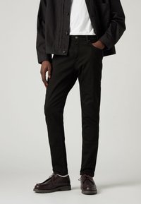 Svarta denimjeans med slim fit, featuring fickor och knappstängning. Bärs med en vit t-shirt och svart jacka. Mörka läderskor.