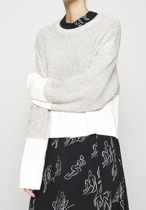 Osoba nosząca luźny, jasnoszary i biały sweter z dzianiny na czarnej sukience z białymi, abstrakcyjnymi rysunkami linii figuralnych.