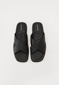 Sorte svarte sandaler med kryssremdesign, glatt syntetisk materiale, flat såle og logo preget på fotsengen.