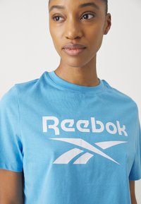 Lyseblå bomulls-t-skjorte med rund hals og korte ermer, med et stort hvitt Reebok-logo grafikk foran.