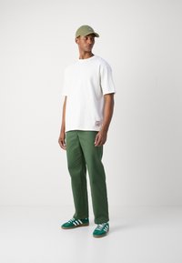 Camiseta blanca de manga corta con un parche colorido, combinada con pantalones verdes y zapatillas verdes con rayas blancas. Gorra oliva.