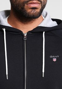 Felpa nera con zip e cappuccio grigio all'interno, con lacci bianchi e zip argentata. Presenta un logo GANT sul petto.