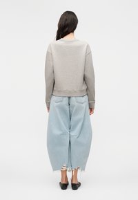 Mujer con cabello largo y oscuro, que lleva una sudadera gris claro, pantalones vaqueros anchos azul claro desgastados y zapatos negros sin cordones, de pie de espaldas.