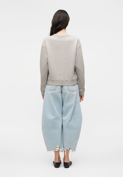 Γυναίκα με μακριά σκουρόχρωμα μαλλιά που φοράει ανοιχτογκρι φούτερ, φαρδιά ανοιχτομπλε distressed τζιν παντελόνια και μαύρα παντόφλες slip-on, με την πλάτη στραμμένη.