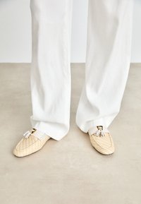 Mocassini beige intrecciati con un motivo a righe, dotati di una fascia in pelle bianca e dettaglio con nappine, abbinati a pantaloni bianchi larghi.