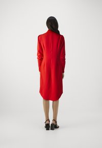 Rotes Satin-Kleid mit langen Ärmeln, hohem Kragen und geschwungener Saumlinie. Gestylt mit schwarzen Absätzen. Die Rückansicht zeigt eine glatte Textur.