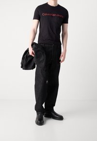 Černé tričko s červeným logem "Calvin Klein Jeans", v kombinaci s tmavými džíny a černými koženými kotníkovými botami, držící černou bundu.