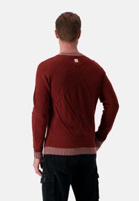 Roter Strickpullover mit strukturierten Rautenmuster, gestreiftem Kragen und Bündchen, rechteckigem Etikett auf der Rückseite, kombiniert mit schwarzen Cordhosen.