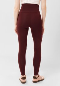 Leggings bordeaux a vita alta con una texture liscia, design aderente e taglio alla caviglia, abbinati a sneakers rosa chiaro.