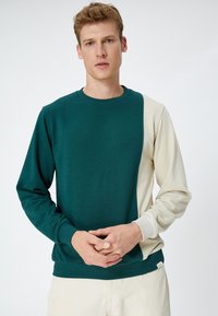 Sudadera en verde oscuro y crema, fabricada con material suave. Cuenta con un cuello redondo, mangas largas y un diseño de bloques de color en contraste.