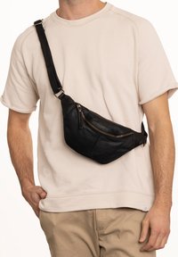 Černá kožená taška přes rameno s zakřiveným designem, přední kapsou na zip a nastavitelným popruhem, nošená přes tělo na béžové košili.