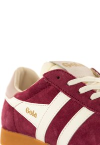 Red suede sneaker with white stripes, cream accents, and gold logo. Features round laces and textured material. Rubber outsole visible.