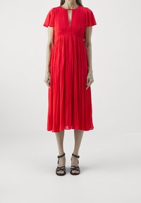 Abito rosso plissettato con maniche a farfalla corte, scollatura a V e vita impero, abbinato a sandali neri con cinturini.