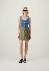 Γυναίκα που φορά μαύρα γυαλιά ηλίου, αμάνικο μπλε τζιν πουκάμισο με κουμπιά, ελαιώδη φούστα τύπου περιτύλιξης και μαύρες ανοιχτές σαγιονάρες, που στέκεται σε απλό φόντο.