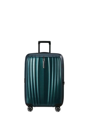 Valigia rigida verde petrolio scuro con manico telescopico e quattro ruote girevoli, in piedi su sfondo bianco.