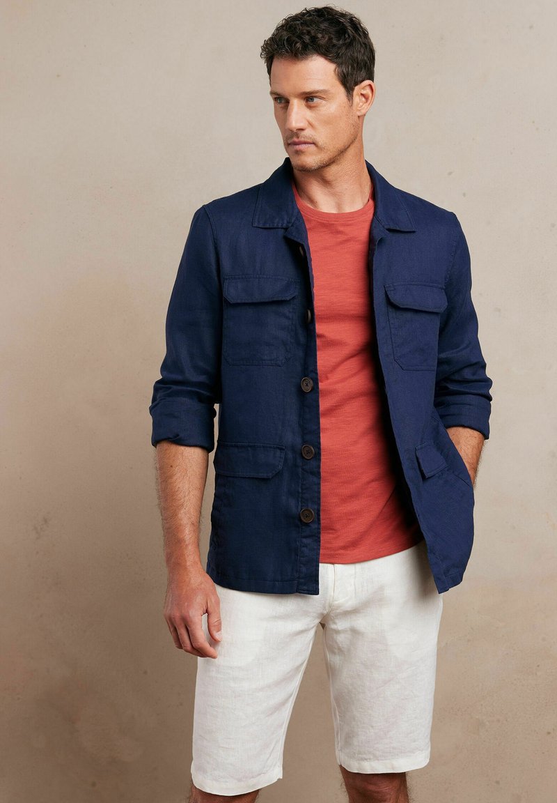 Granatowa lniana kurtka z czterema przednimi kieszeniami, zapinana na guziki, z podwiniętymi rękawami. Nosi się ją na koralowej koszulce i w połączeniu z białymi szortami.