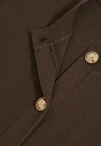 Rudas megzta palaidinė su V formos iškirpte, su auksiniais mygtukais ir įmaniškais rankogaliais. Lygus audinys su švariu, klasikiniu stiliumi.