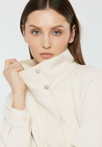 Woman with light skin and blue eyes wearing a cream-colored high-collar jacket, holding the collar with one hand, neutral expression.