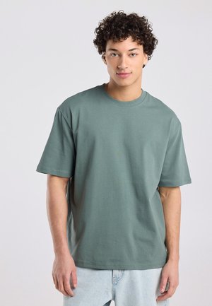 Jonge persoon met krullend haar, gekleed in een loszittend groen T-shirt met korte mouwen en lichtblauwe spijkerbroek, staand tegen een effen lichte achtergrond.