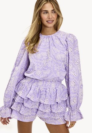 Mujer que lleva una blusa de encaje floral color lila claro con mangas largas abullonadas y pantalones cortos de volantes en capas a juego, sonriendo y mirando de lado.