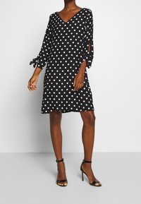 Nainen, joka käyttää mustaa polven pituista mekkoa valkoisilla pilkuilla, kolmivartisia hihoja sidontatiedoilla ja mustia korkokenkiä.