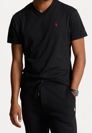 Schwarzes Baumwoll-V-Ausschnitt-T-Shirt mit kurzen Ärmeln, mit einem kleinen roten Logo auf der Brust. Getragen mit schwarzen Hosen und einer silbernen Uhr.