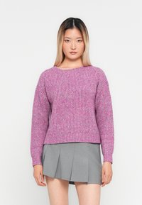 Donna che indossa un maglione lavorato a maglia rosa e una gonna plissettata grigia, in piedi davanti a uno sfondo bianco uniforme.