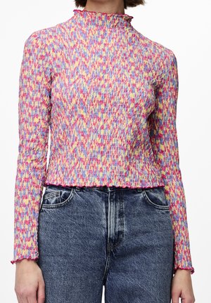 Kvinde iført en langærmet, flerfarvet smocked bluse med flæsekanter, kombineret med højttaljede, falmede blå jeans.