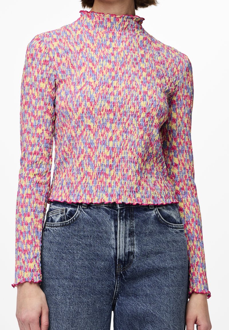 Femme portant un chemisier smocké multicolore à manches longues avec des bords volantés, associé à un jean bleu délavé taille haute.