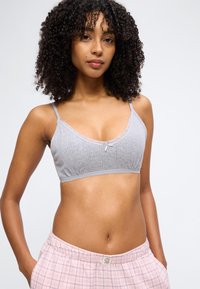 Νεαρή γυναίκα με σγουρά μαλλιά που φοράει γκρι πλεκτό μπραλέτο και ροζ πιτζάμες σε καρό σχέδιο, στέκεται με τα χέρια στις τσέπες.