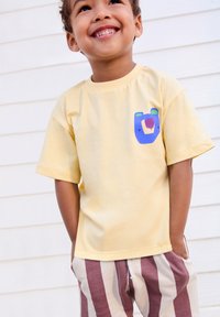 Yellow cotton t-shirt with short sleeves featuring a colorful bear graphic on the chest, paired with striped beige and maroon shorts.