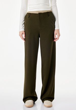 Persoon draagt olijfgroene wijde broek, beige schoenen en een wit top met lange mouwen tegen een effen witte achtergrond.