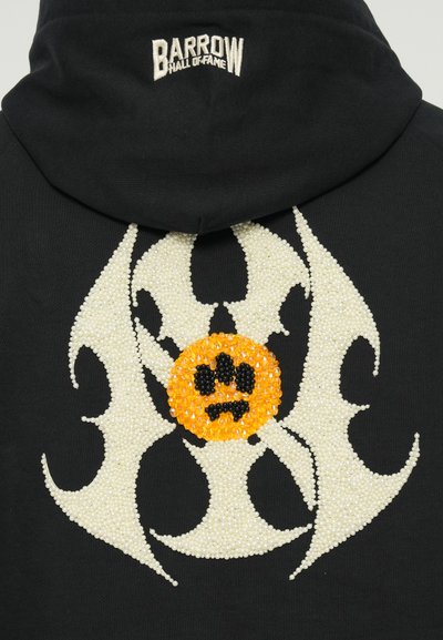 Čierna mikina s kapucňou, ktorá má biele koráliky usporiadané do vírivého vzoru, so stredovým oranžovým kruhovým motívom a čiernou stopou zvieraťa.