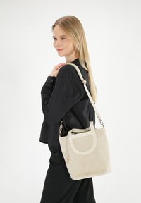 Beige ruskind håndtaske med en struktureret form, med en front cut-out detalje, justerbar skulderrem og hardware i guld tone.