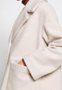 Abrigo texturizado de color beige claro con cuello con muesca, corte oversize y bolsillos laterales, que muestra un material suave y aterciopelado.