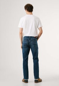 Jeans de mezclilla azul oscuro con corte recto, dos bolsillos traseros y una etiqueta de cuero en la pretina. Combinados con una camiseta blanca de cuello redondo.