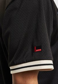 Tricou sport negru cu mânecă scurtă, dintr-un material mesh texturat, având un manșon cu dungi albe și negre și un mic patch cu logo pe manecă.